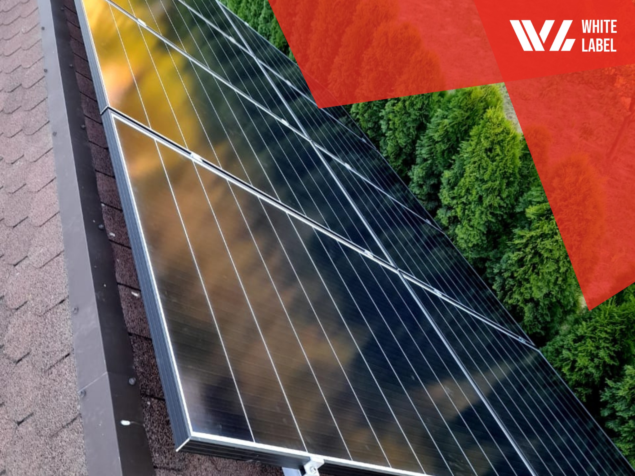 Panele słoneczne na dachu pokrytym brązową dachówką bitumiczną, z widocznym logo firmy w prawym górnym rogu i odbiciem drzew w panelach.