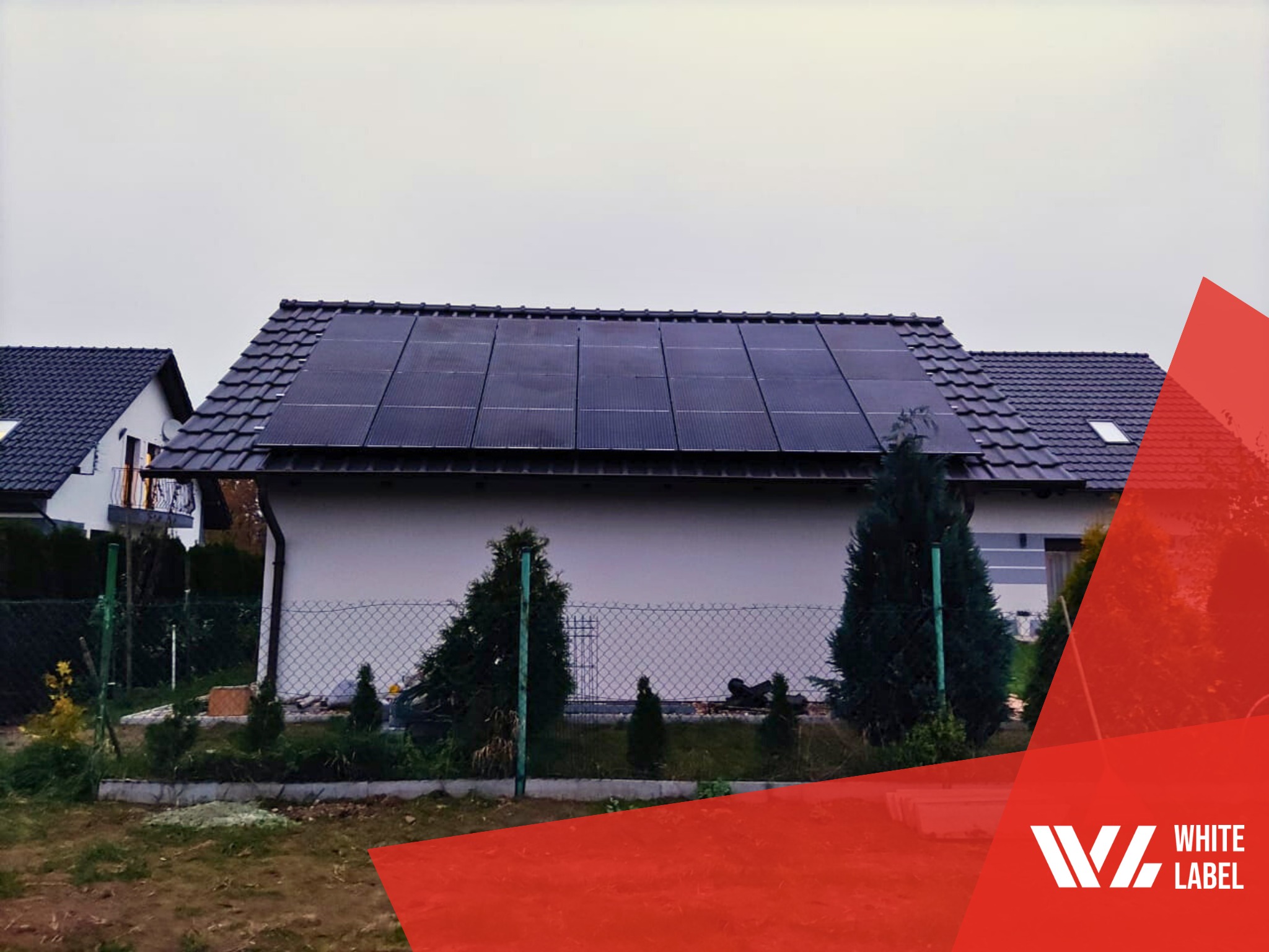 Dom jednorodzinny z panelami słonecznymi na dachu, widok z ogrodu, szara pogoda, dolny róg zdjęcia zasłonięty czerwonym logotypem.