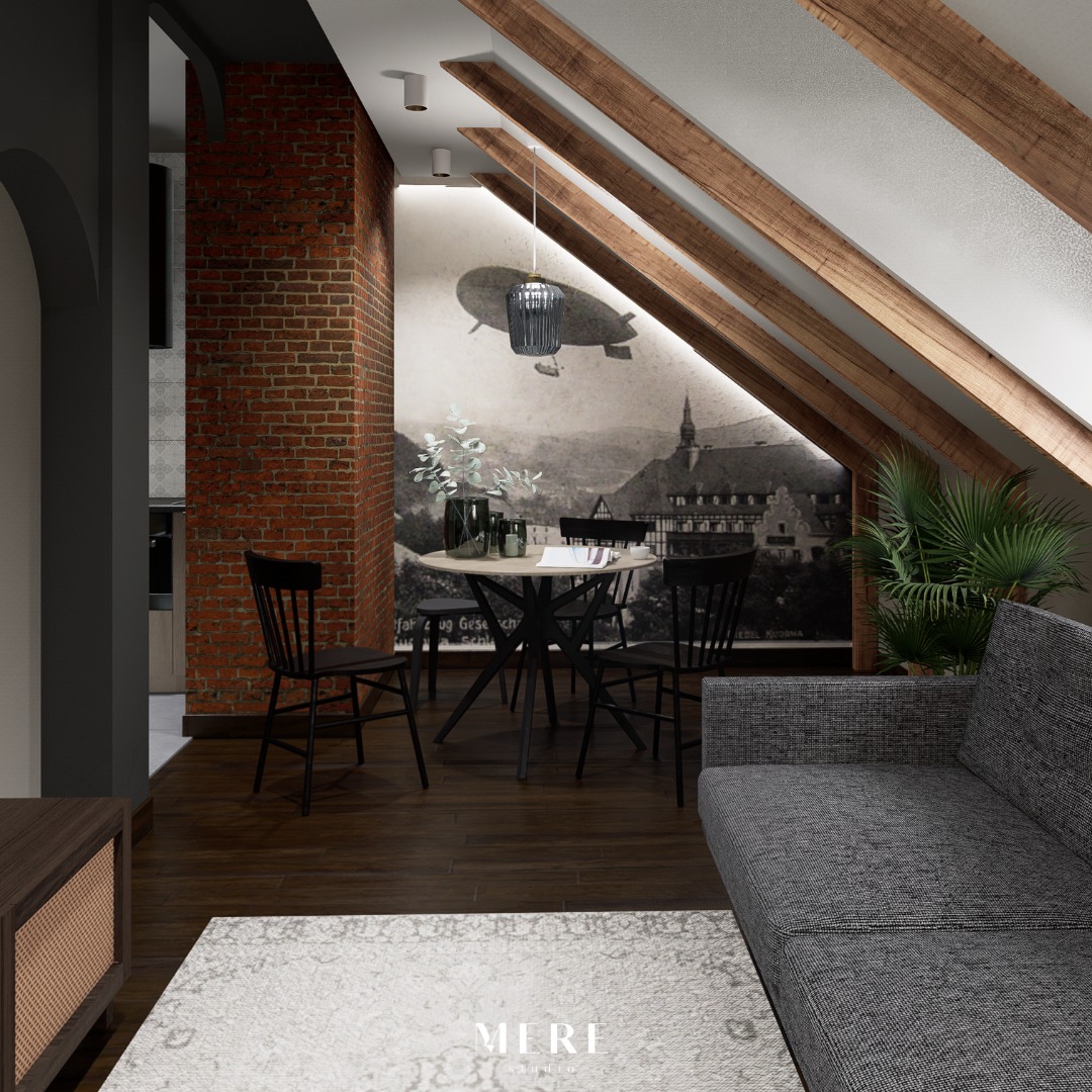 Wnętrze poddasza ze skośnym sufitem z drewnianymi belkami, ceglaną ścianą i fototapetą z motywem sterowca nad okrągłym stołem z czarnymi krzesłami, szara sofa w tle.