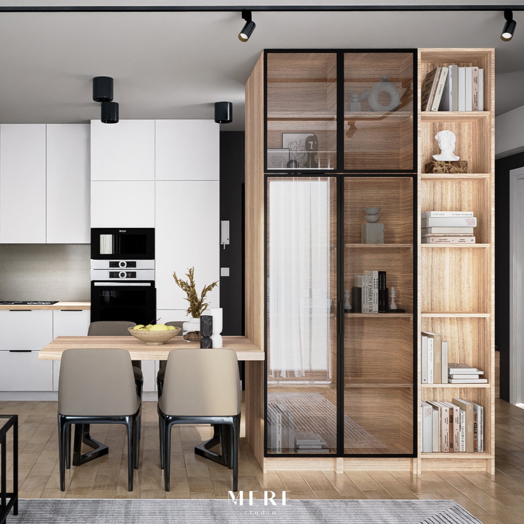 Nowoczesna kuchnia z białymi szafkami, drewnianym stołem i krzesłami w kolorze taupe, oraz regałem z przeszklonymi drzwiami i otwartymi półkami, oświetlona czarnymi reflektorami punktowymi.