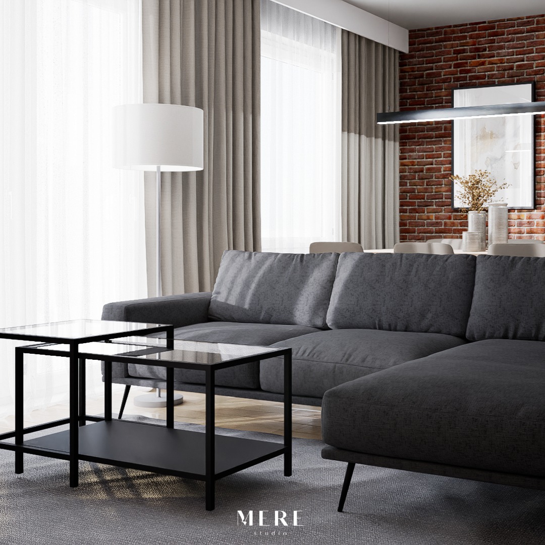 Nowoczesny salon z szarą sofą narożną, stolikami kawowymi z czarnymi ramami i szklanymi blatami, oświetlony lampą podłogową i wiszącą lampą liniową, z ceglaną ścianą w tle i zasłonami w kolorze taupe.
