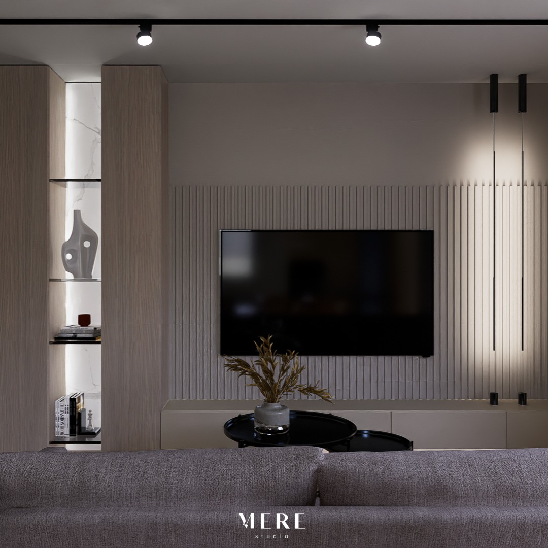 Salon z telewizorem na ścianie z dekoracyjnymi pionowymi panelami, oświetleniem punktowym i podłużnymi lampami wiszącymi, wazon z suchymi roślinami na czarnym stoliku kawowym, widoczny fragment...
