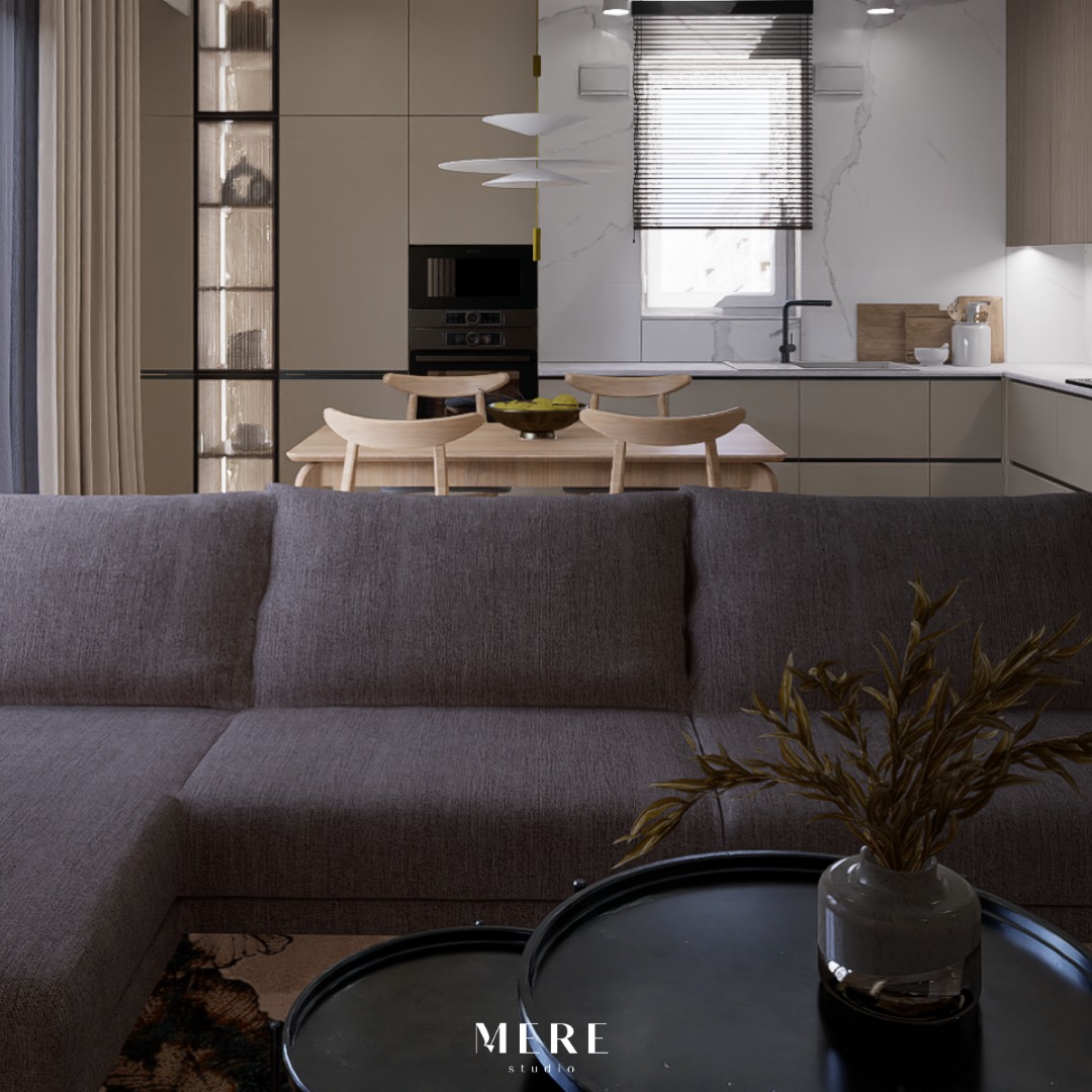 Wnętrze salonu z aneksem kuchennym w neutralnych barwach, z widoczną szarą sofą, stolikami kawowymi z dekoracyjną rośliną w wazonie oraz stołem z krzesłami w tle.