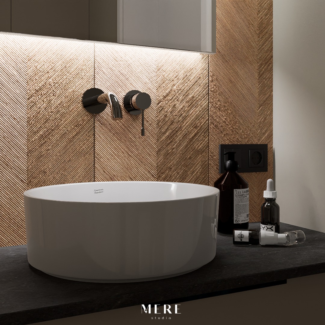 Nowoczesna łazienka z okrągłą, białą umywalką nablatową American Standard, czarną armaturą ścienną na tle ściany wykończonej płytkami w jodełkę w odcieniach brązu i beżu, uzupełniona o czarny blat...