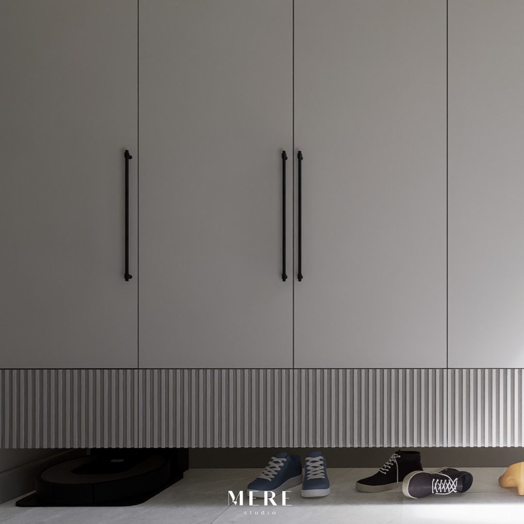 Szare szafy wnękowe z długimi, czarnymi uchwytami, dolna część wykończona dekoracyjnymi pionowymi panelami, pod nimi kilka par butów i robot sprzątający.