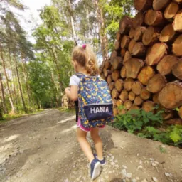 Dziewczynka z blond włosami idzie leśną ścieżką, na plecach ma plecak z nadrukiem i imieniem 'HANIA', obok stos drewna.