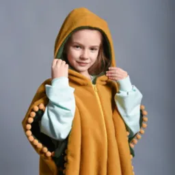 Dziewczynka w musztardowym, pluszowym ponczo z kapturem i pomponami, zapinanym na zamek, prezentuje strój wykonany w Wołowie.