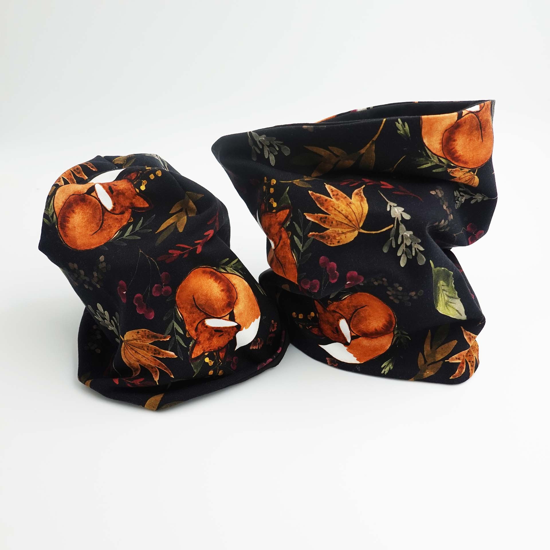 Dwa kominy uszyte z czarnej dzianiny w rudego lisa i jesienne liście, leżące na białym tle.