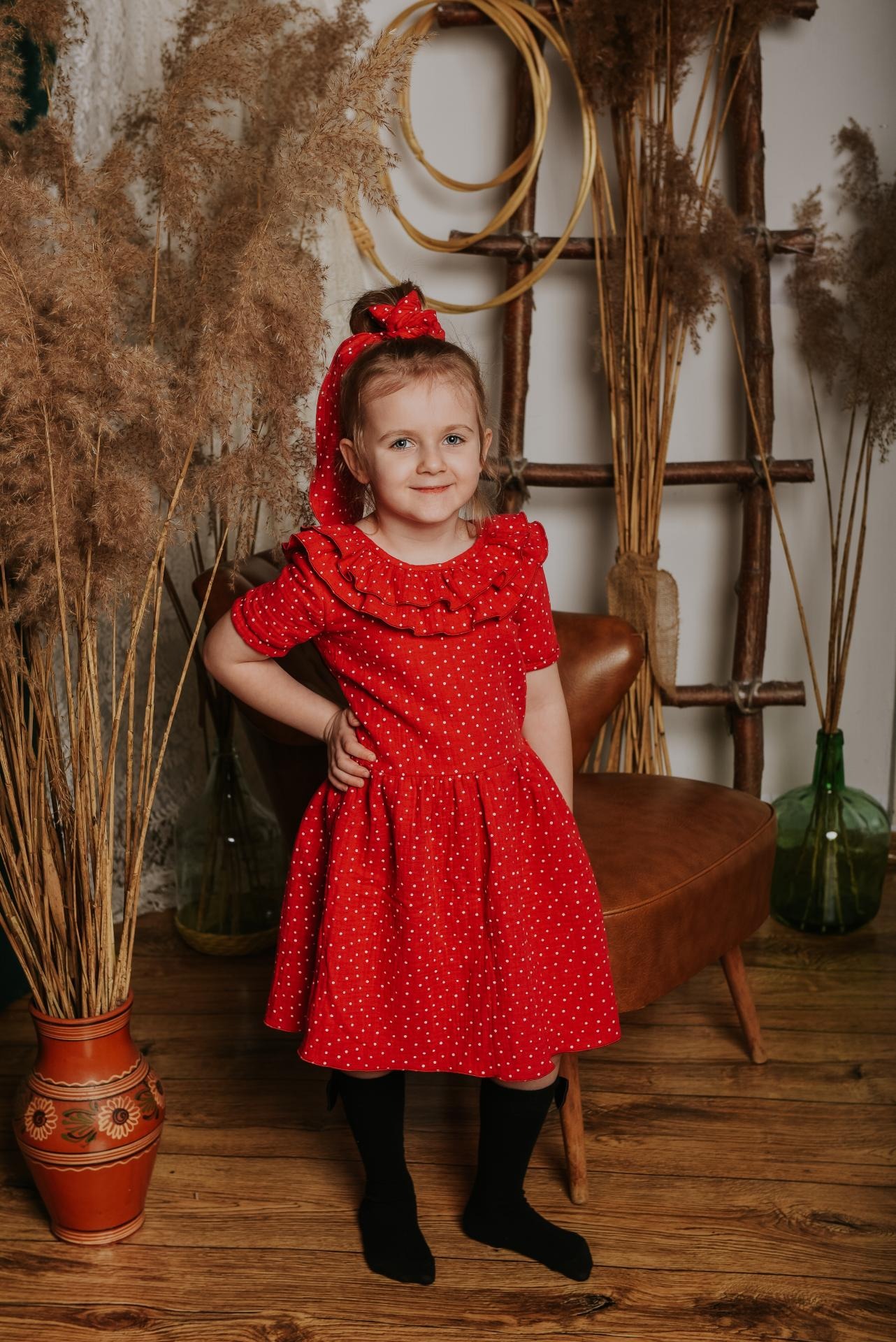Uśmiechnięta dziewczynka w czerwonej sukience w białe kropki z kokardą, pozująca na tle drewnianej drabiny i dekoracyjnych traw w wazonie.