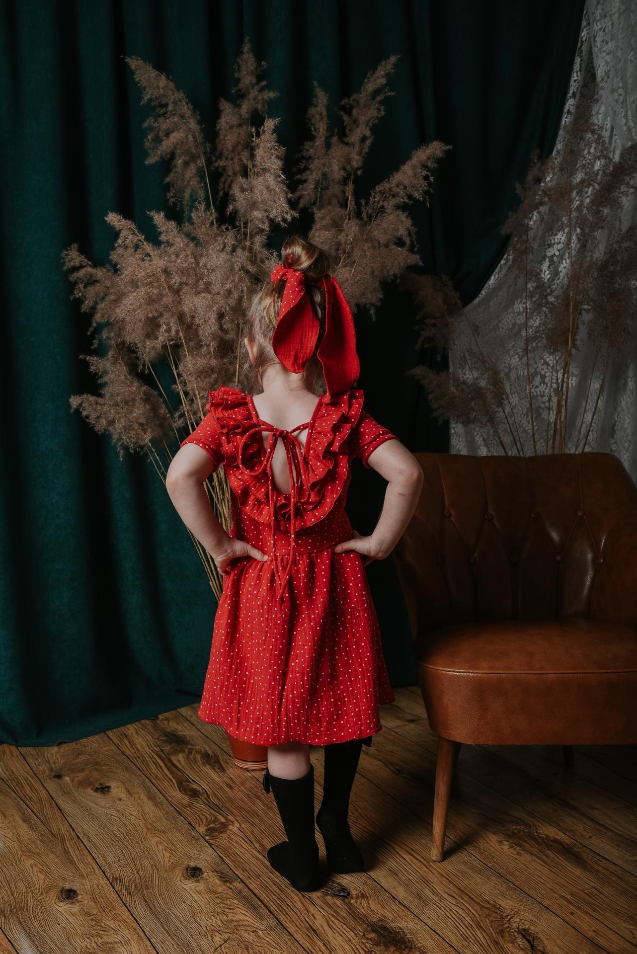 Dziewczynka tyłem w czerwonej sukience w białe kropki z falbankami i kokardą, ręce na biodrach, na tle zielonej zasłony i pampasów.