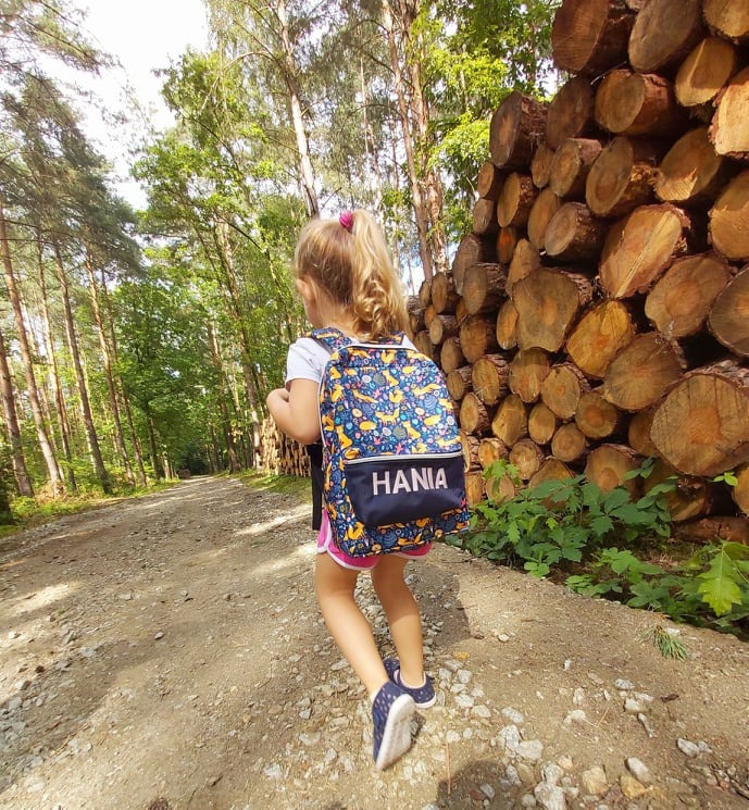 Dziewczynka z blond włosami idzie leśną ścieżką, na plecach ma plecak z nadrukiem i imieniem 'HANIA', obok stos drewna.