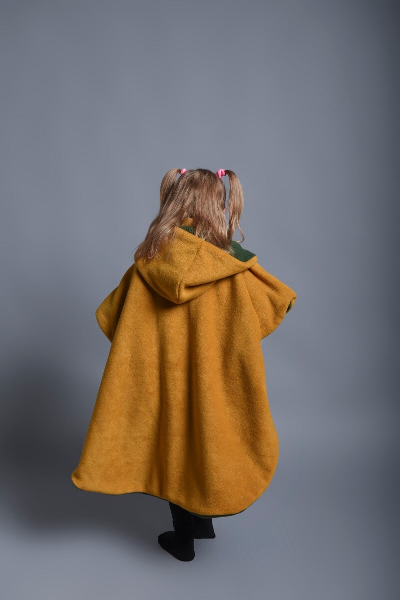 Dziewczynka z blond włosami spiętymi w dwa kucyki z różowymi gumkami, ubrana w musztardowy, polarowy płaszcz z kapturem i zieloną podszewką, stojąca tyłem na szarym tle.