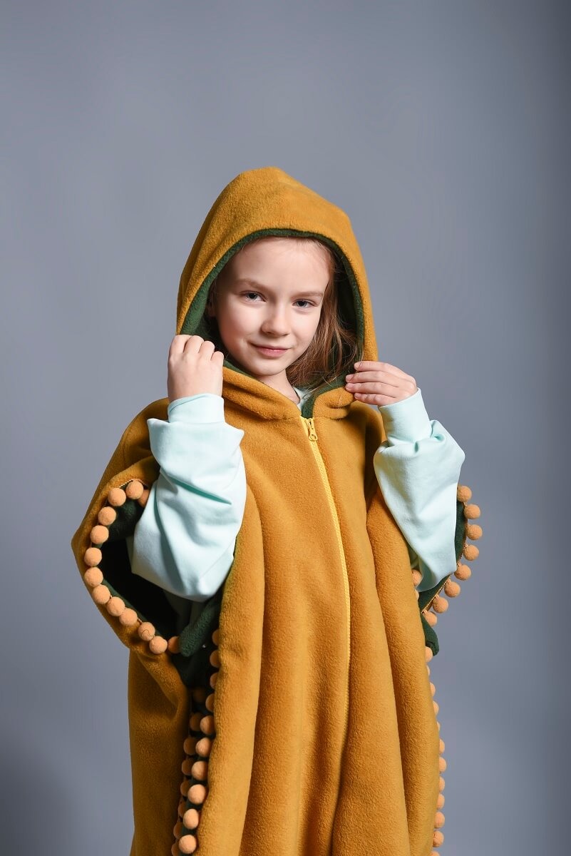 Dziewczynka w musztardowym, pluszowym ponczo z kapturem i pomponami, zapinanym na zamek, prezentuje strój wykonany w Wołowie.