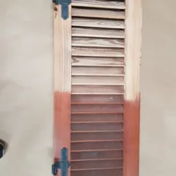 Drewniana okiennica żaluzjowa, częściowo odnowiona, z widoczną różnicą w kolorze drewna przed i po renowacji, z zamontowanymi czarnymi zawiasami.