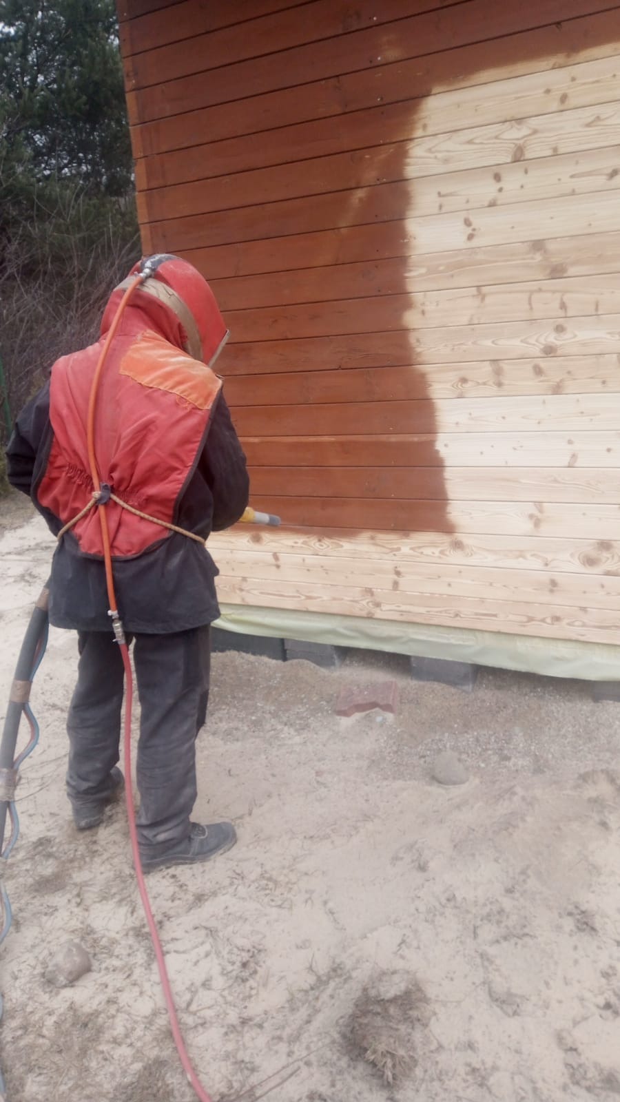 Pracownik w kombinezonie ochronnym czyści drewnianą elewację budynku, widoczny kontrast między oczyszczoną a surową powierzchnią drewna, na ziemi piasek.