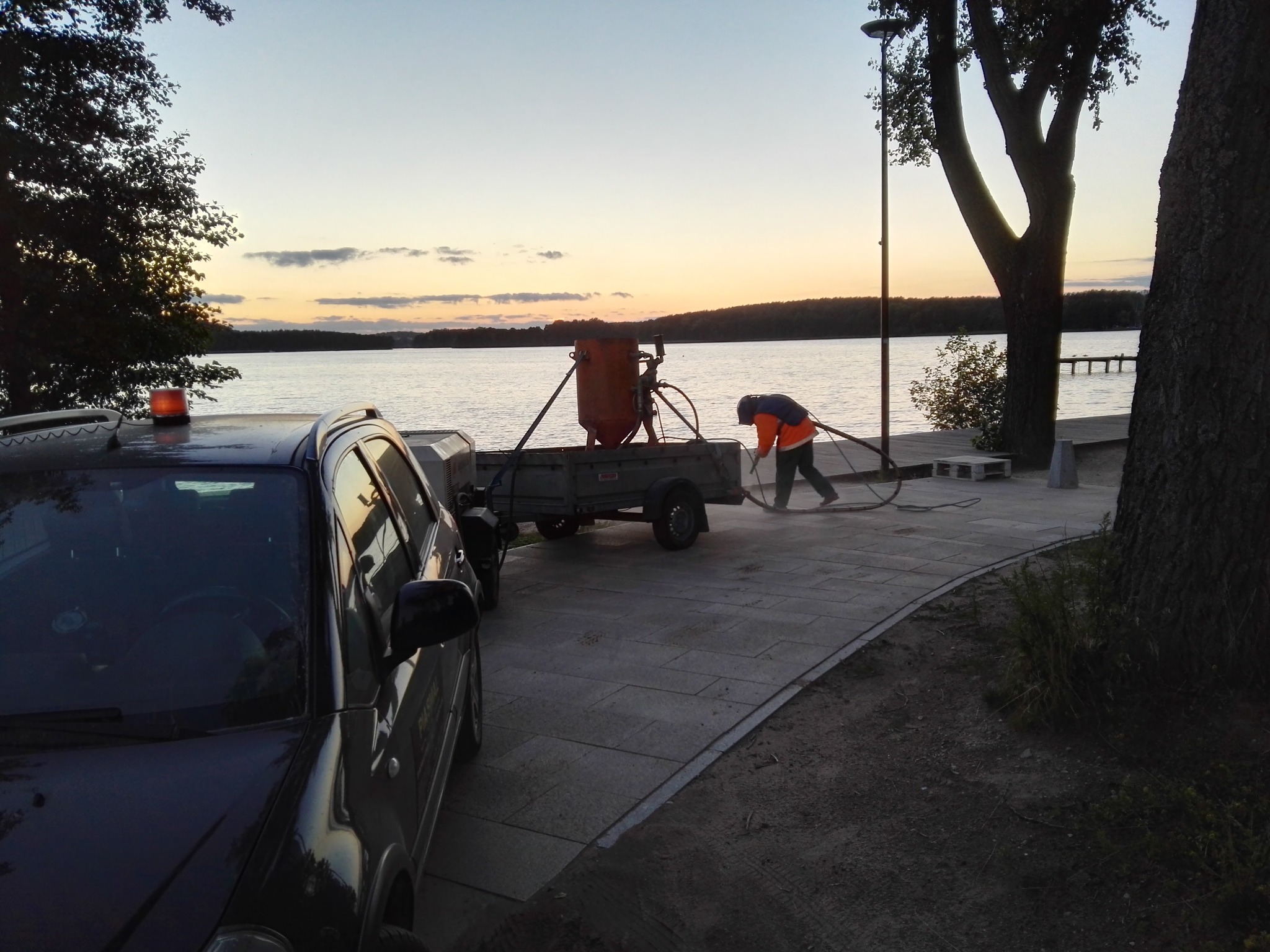 Pracownik w kamizelce odblaskowej czyści chodnik z kostki brukowej za pomocą urządzenia do piaskowania, które stoi na przyczepce za samochodem, w tle jezioro i zachodzące słońce.