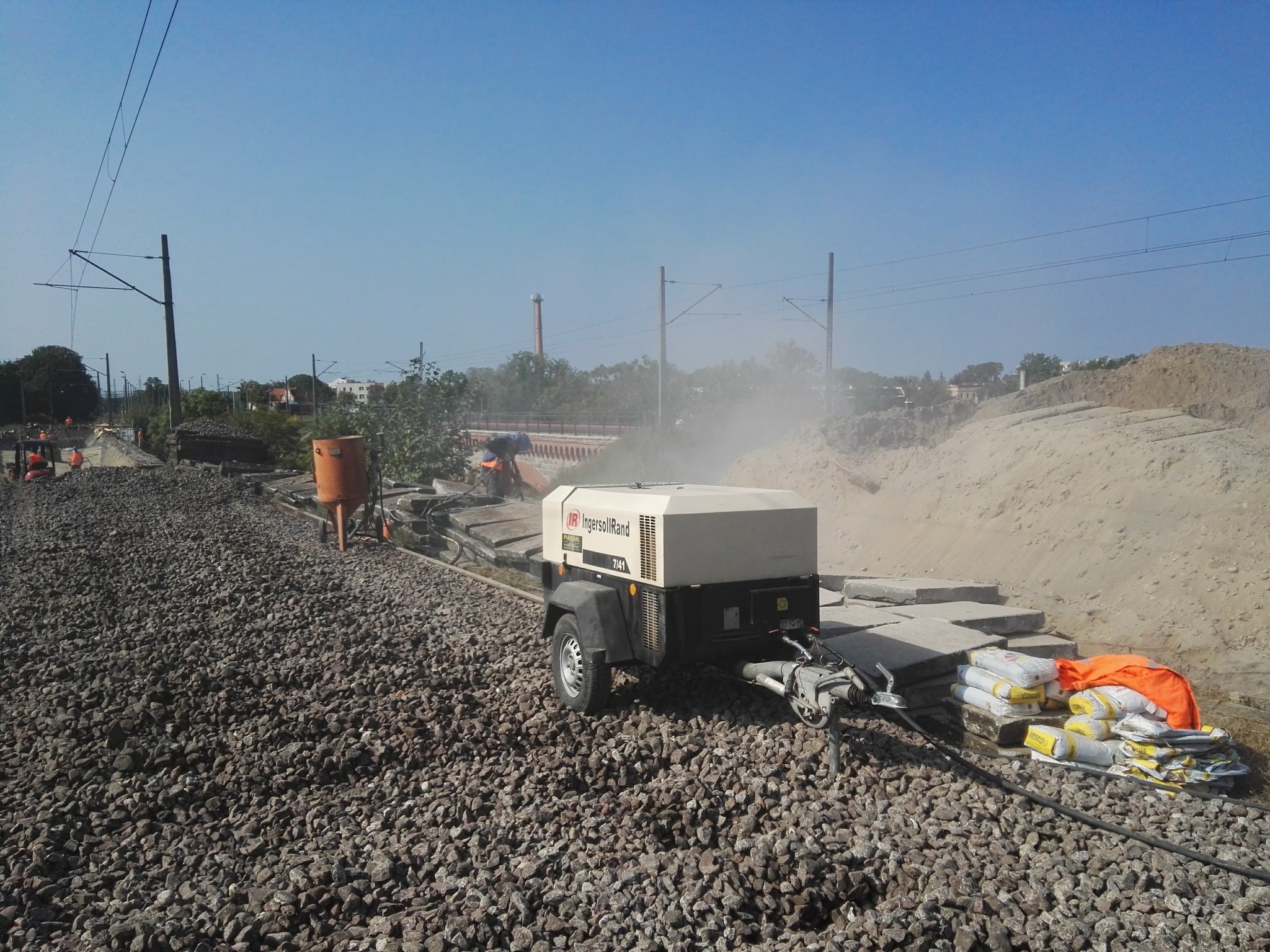Mobilna piaskarka Ingersoll Rand 7/41 na torowisku kolejowym w trakcie pracy, unoszący się pył, w tle widoczne elementy infrastruktury kolejowej.