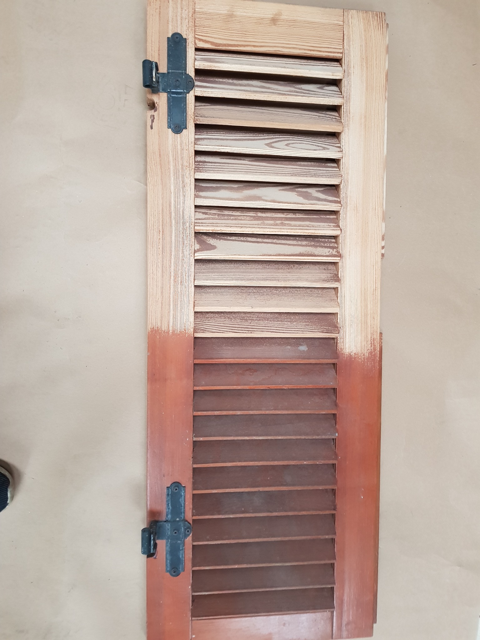 Drewniana okiennica żaluzjowa, częściowo odnowiona, z widoczną różnicą w kolorze drewna przed i po renowacji, z zamontowanymi czarnymi zawiasami.
