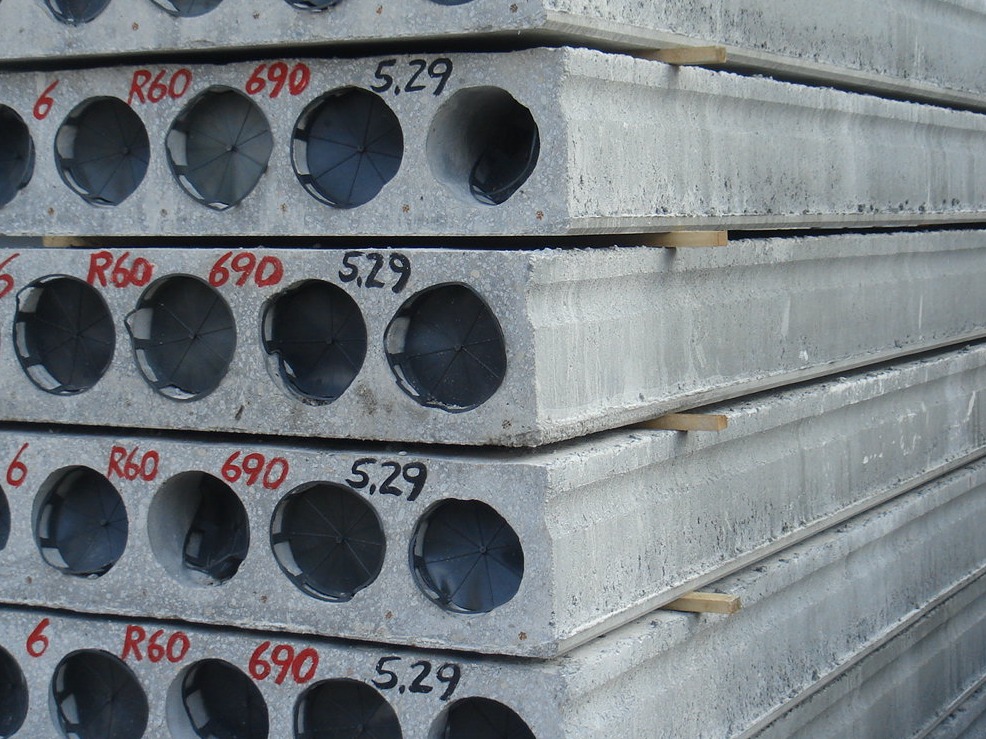 Szare, żelbetowe płyty stropowe z okrągłymi otworami, oznaczone numerami 'R60 690 5.29', składowane w Białe Błota i oddzielone drewnianymi przekładkami.