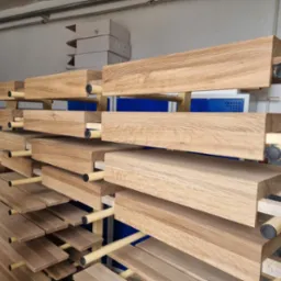 Drewniane stopnie schodowe ułożone na metalowym regale w warsztacie, widoczne puszki z farbą w tle.