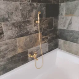 Biała wanna z baterią i deszczownicą w kolorze złotym, obłożona płytkami imitującymi kamień w odcieniach szarości i brązu.