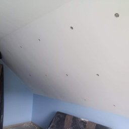 Izobud - Poddasze z ociepleniem wełną mineralną, widoczne otwory wentylacyjne w suficie i maszyna do wdmuchiwania izolacji, ściany pomalowane na niebiesko.