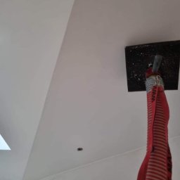 Izobud - Biały sufit poddasza ze skosem, widoczne okno dachowe, otwory po lampach oraz narzędzie do szpachlowania z czerwoną rurą doprowadzającą masę szpachlową.