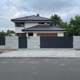Nowoczesna posesja z szarą, poziomą bramą lamelową i betonowym ogrodzeniem modułowym na tle domu z ciemną dachówką i szarą kostką brukową podjazdu.