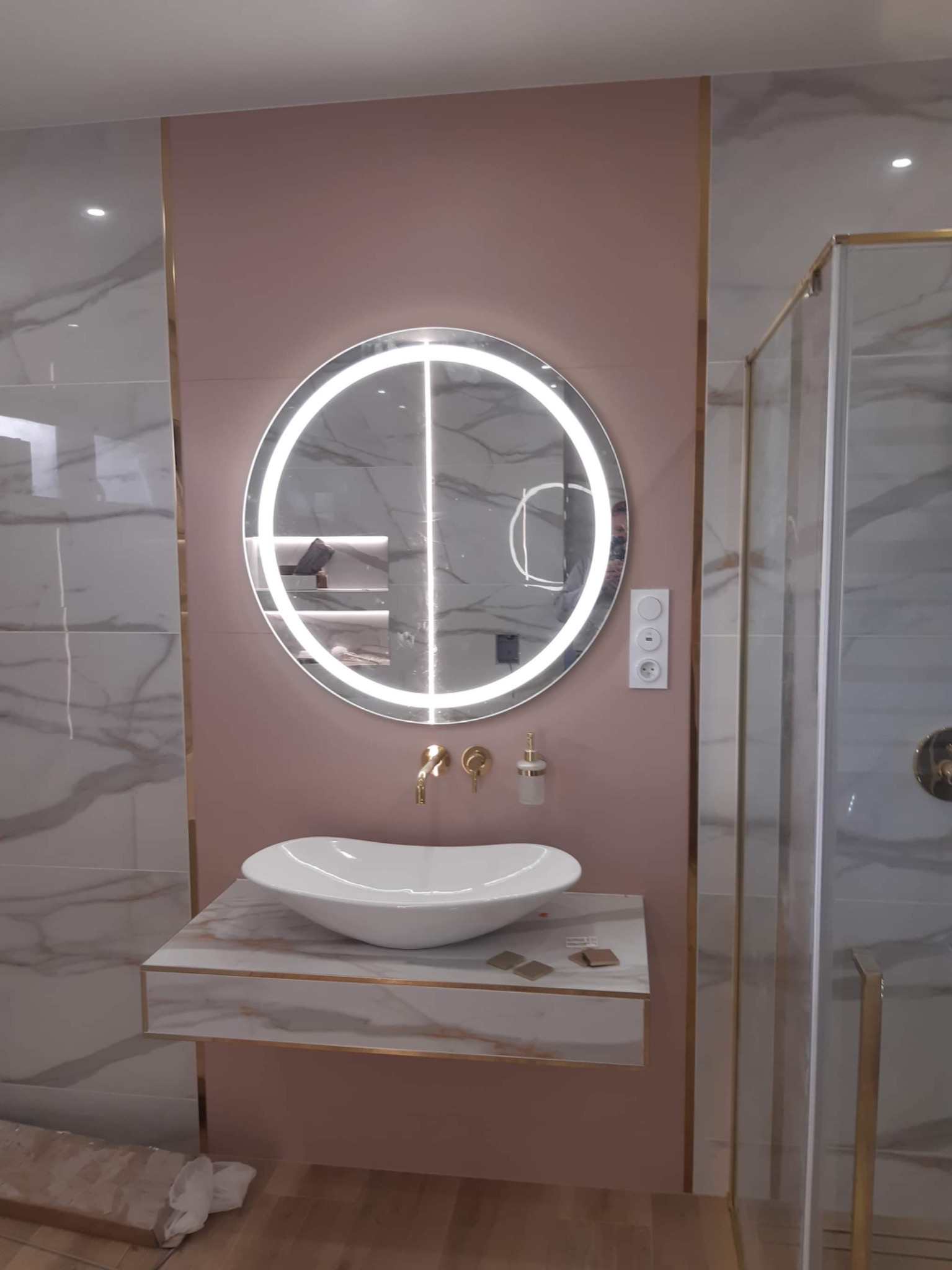 Nowoczesna łazienka z okrągłym lustrem LED, umywalką nablatową na marmurowym blacie z złotymi akcentami, różową ścianą i kabiną prysznicową z elementami w kolorze złota.