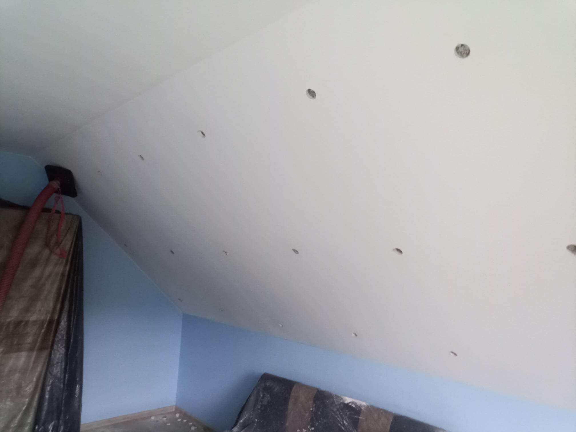 Poddasze z ociepleniem wełną mineralną, widoczne otwory wentylacyjne w suficie i maszyna do wdmuchiwania izolacji, ściany pomalowane na niebiesko.