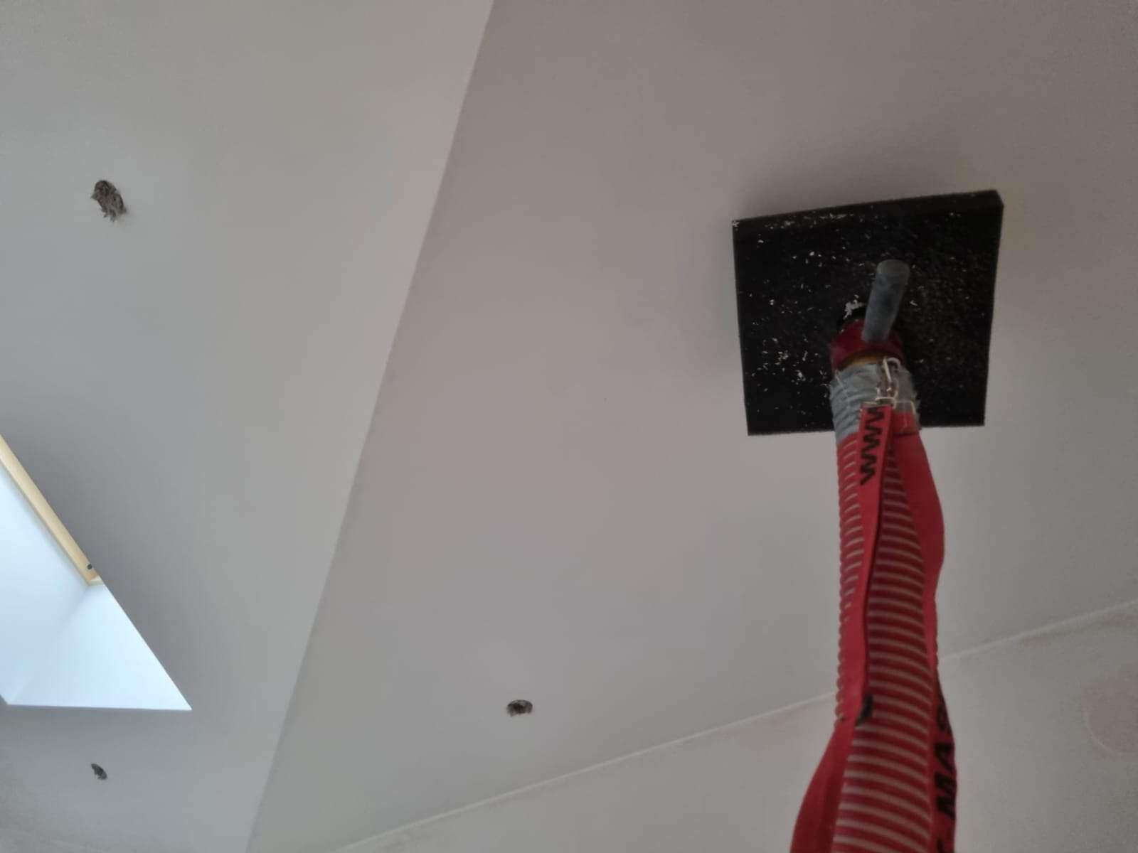 Biały sufit poddasza ze skosem, widoczne okno dachowe, otwory po lampach oraz narzędzie do szpachlowania z czerwoną rurą doprowadzającą masę szpachlową.