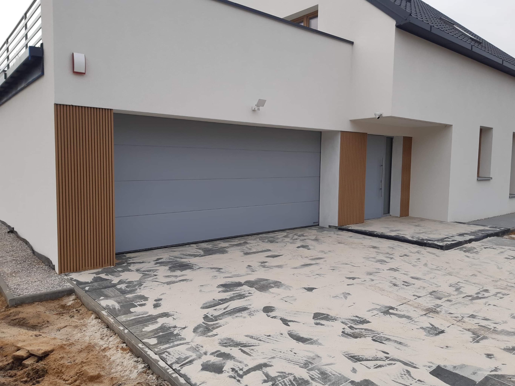 Nowoczesny dom z szarą bramą garażową segmentową i drewnianą okładziną elewacyjną, kostka brukowa w trakcie układania, widoczne ślady piasku na powierzchni.
