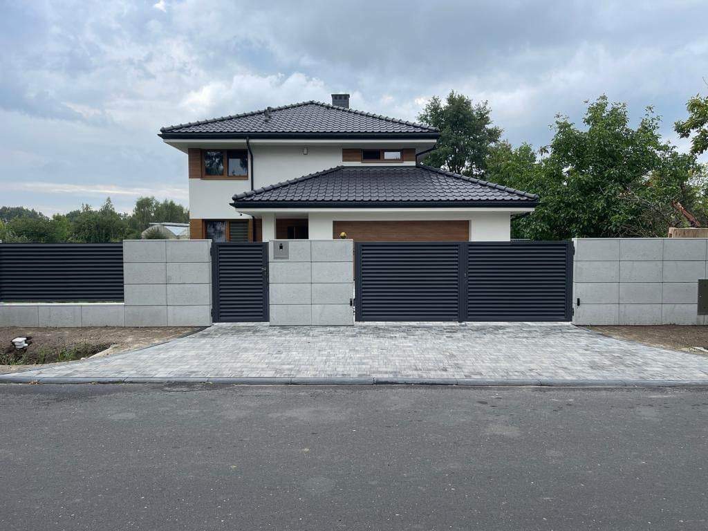 Nowoczesna posesja z szarą, poziomą bramą lamelową i betonowym ogrodzeniem modułowym na tle domu z ciemną dachówką i szarą kostką brukową podjazdu.