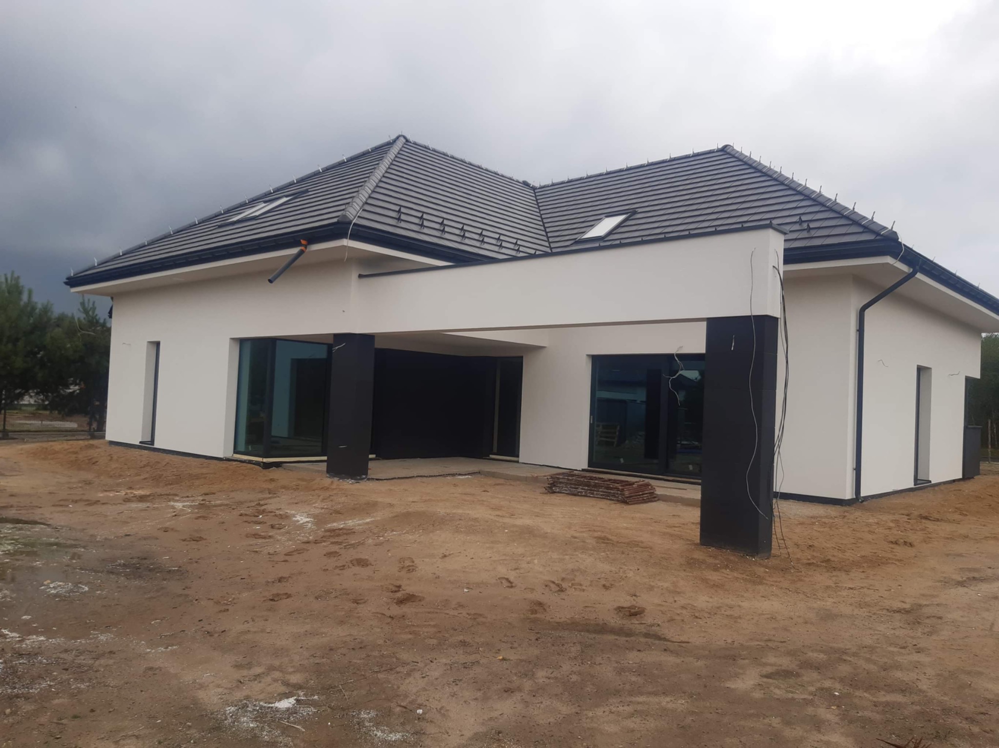 Nowoczesny dom jednorodzinny z białymi ścianami i czarnymi elementami konstrukcyjnymi, szary dach z dachówką, widoczne elementy instalacji elektrycznej na zewnątrz, teren wokół domu w trakcie prac...