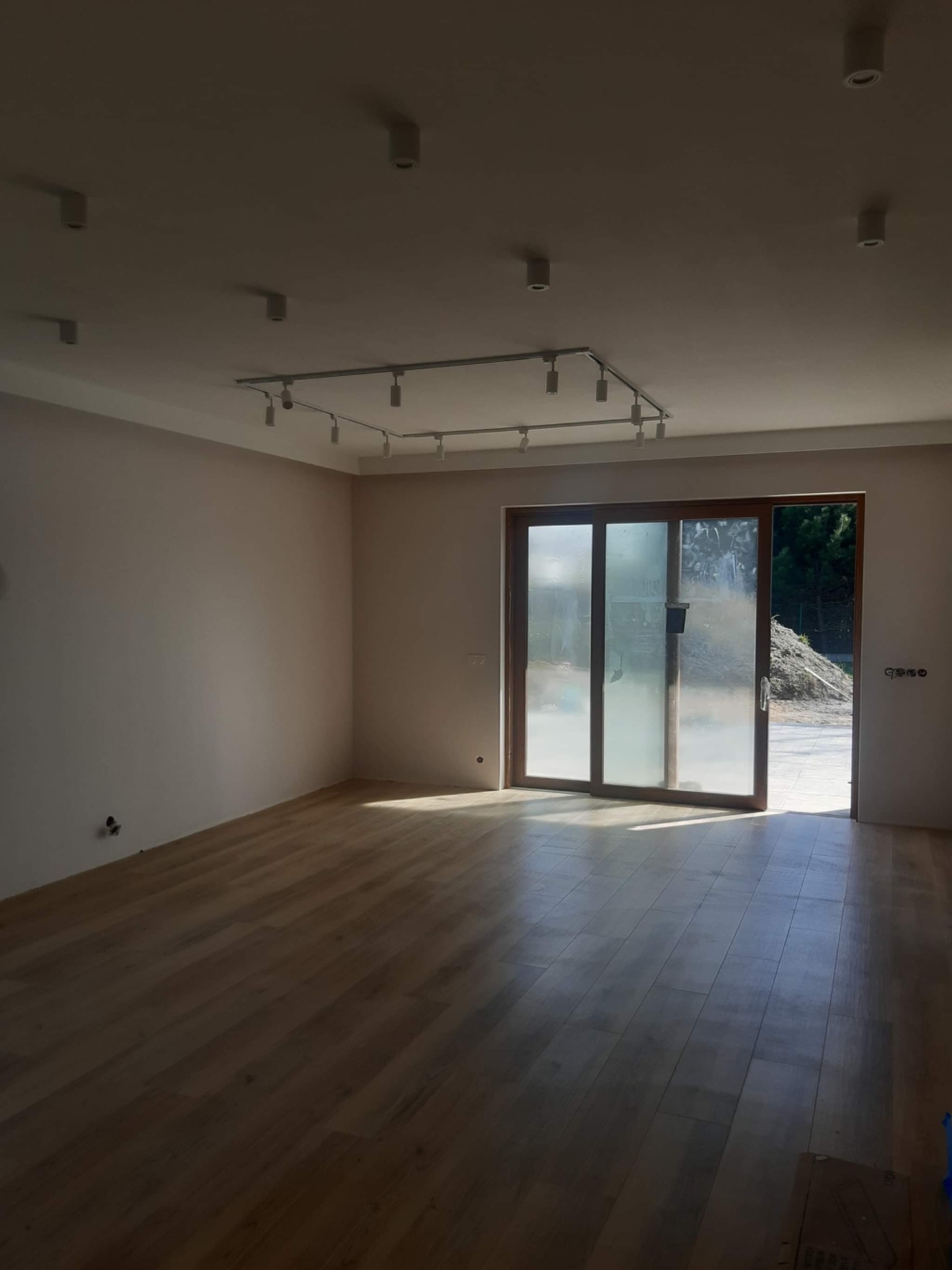 Pusta przestrzeń z nową drewnianą podłogą, świeżo pomalowanymi ścianami w odcieniach beżu i zamontowanym systemem oświetlenia szynowego na suficie, przesuwne drzwi balkonowe z widokiem na zewnątrz.