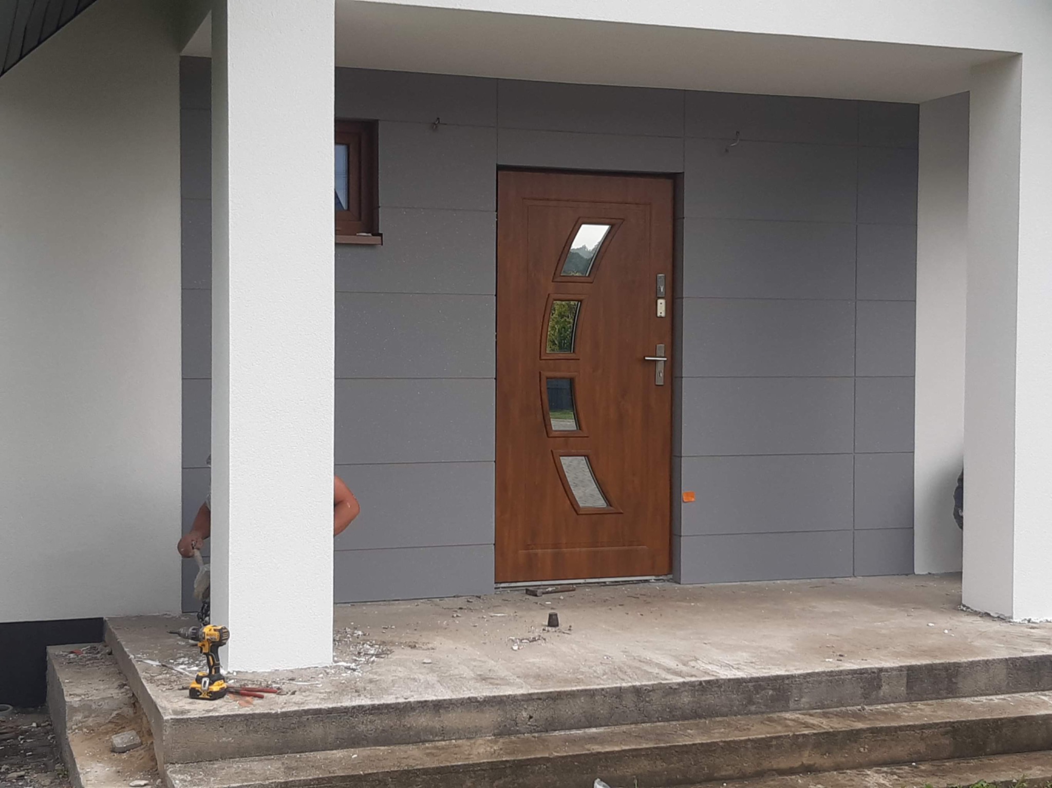 Montaż nowoczesnych, drewnopodobnych drzwi wejściowych z geometrycznymi przeszkleniami w szarej elewacji, widoczne narzędzia i fragment pracownika.