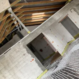 Wnętrze poddasza w trakcie budowy z widocznym systemem ogrzewania podłogowego, rurki ułożone na folii izolacyjnej, ściany z bloczków, drewniana konstrukcja dachu z oknami dachowymi.