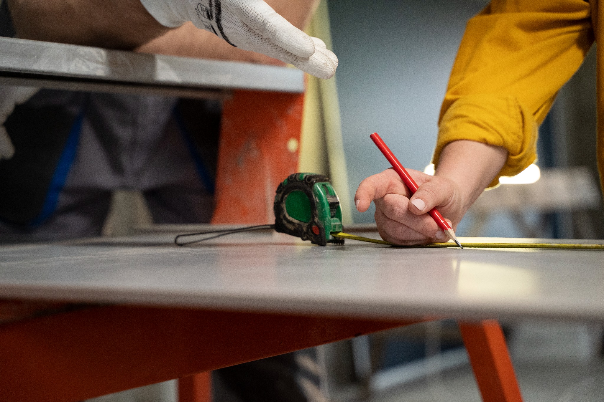 Przygotowanie do nałożenia gładzi: zielona miara zwinięta leży obok czerwonej kredki, którą pracownik w żółtej koszuli zaznacza linię na szarej powierzchni.