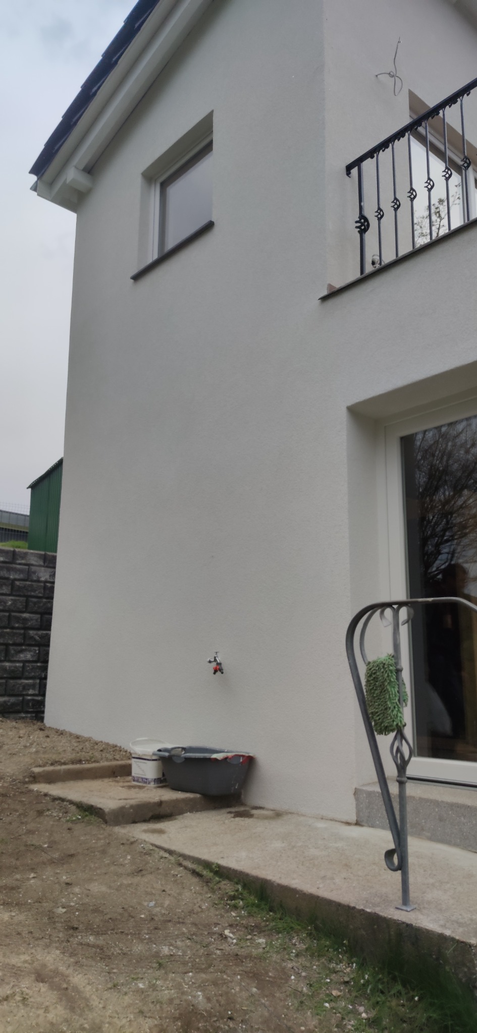 Świeżo pomalowana elewacja domu jednorodzinnego w kolorze białym, widoczny kran na ścianie, wiadro z farbą i szary pojemnik na podłożu, fragment balustrady z zieloną gąbką.