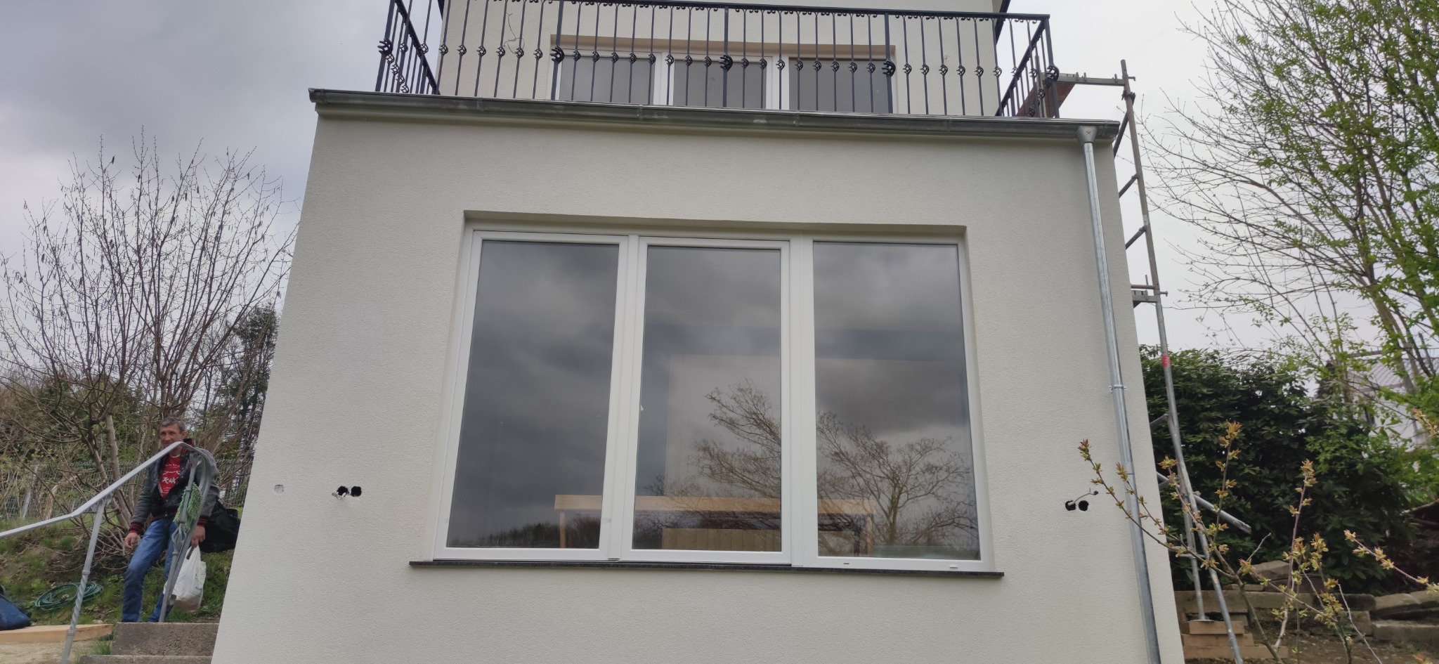 Biała elewacja domu z dużym oknem trzyskrzydłowym, balkonem z kutą balustradą na górze i drabina oparta o ścianę, widoczny mężczyzna schodzący po schodach.