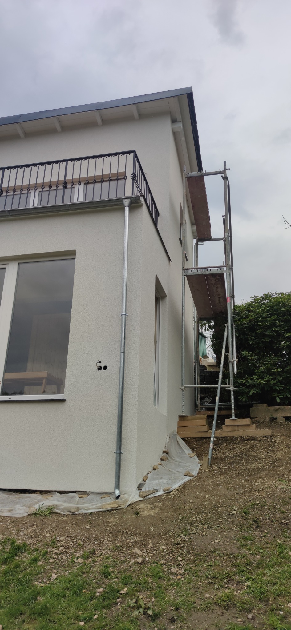 Świeżo pomalowana, jasna elewacja domu jednorodzinnego z balkonem z metalową balustradą, widoczny fragment rusztowania i rynna, teren wokół zabezpieczony folią.
