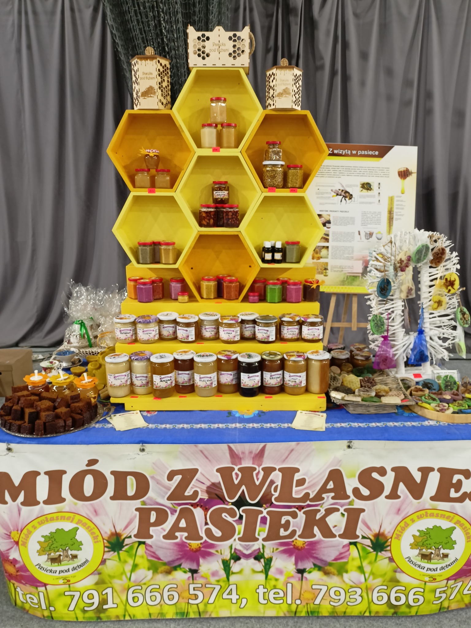 Stoisko z różnymi rodzajami miodów i produktami pszczelimi na targach, z żółtym regałem w kształcie plastra miodu i banerem reklamowym z nazwą pasieki.