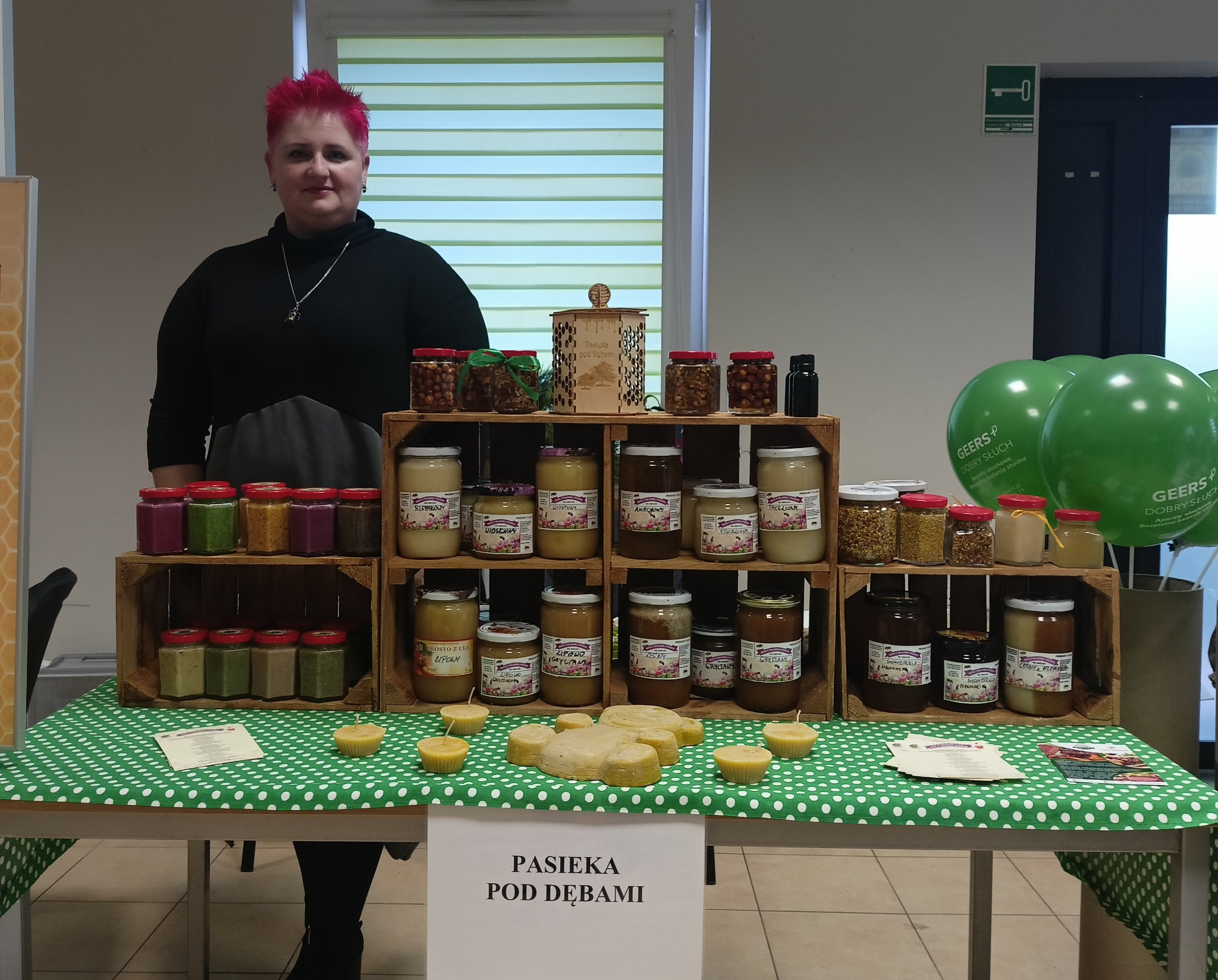 Stoisko z różnymi rodzajami miodów i produktami pszczelimi na zielonym obrusie w kropki, prezentowane przez kobietę z różowymi włosami, w tle balony z logo firmy.