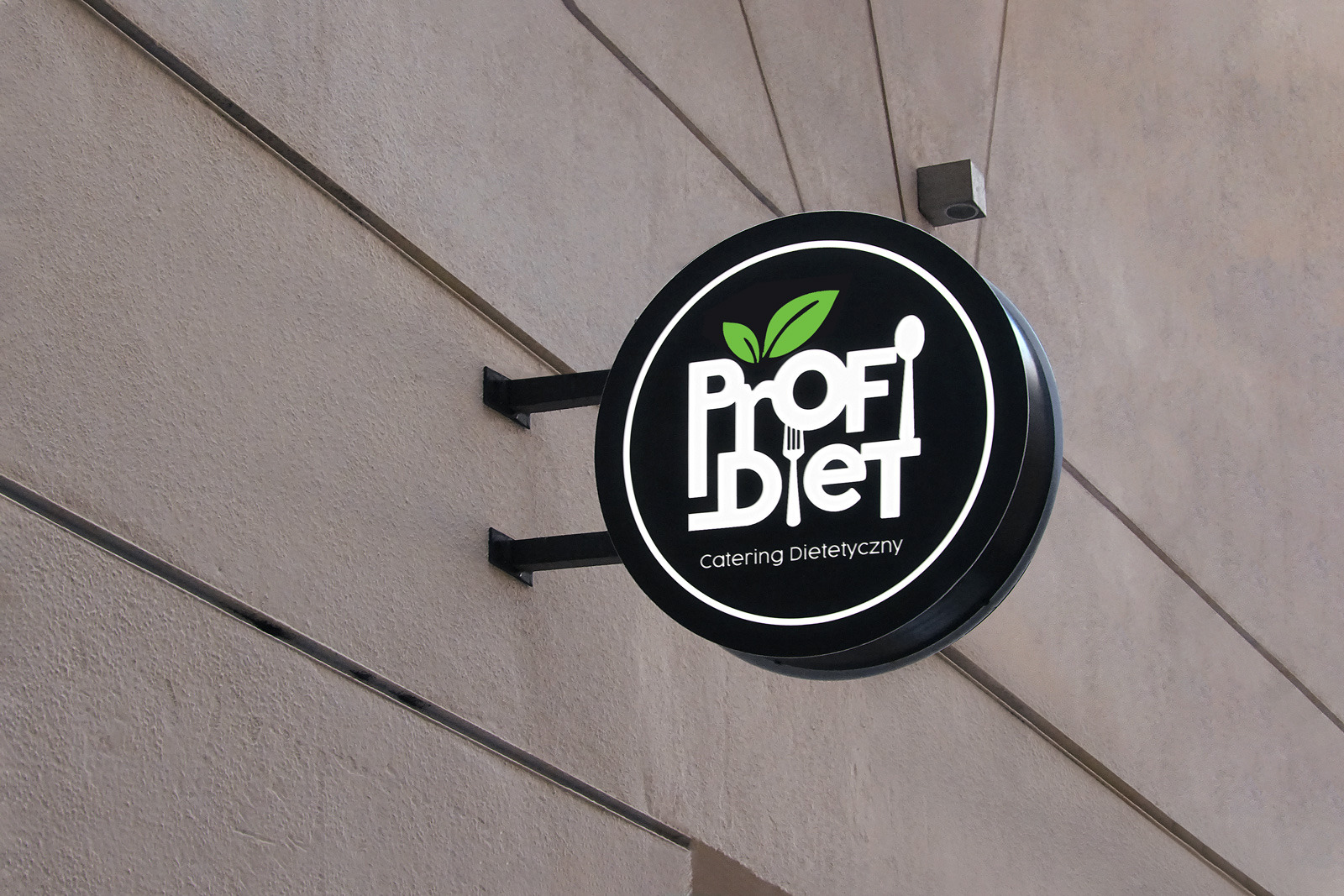 Okrągły szyld reklamowy z logo 'Profi Diet Catering Dietetyczny' umieszczony na elewacji budynku. Logo zawiera zielone liście, łyżkę i widelec.
