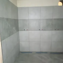 Ściany łazienki wyłożone szarymi płytkami w dwóch odcieniach, z widocznymi przyłączami wodnymi i otworem w górnym rogu ściany.