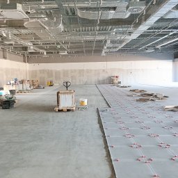 MIŁOSZ TOMKOWICZ - Układanie płytek podłogowych w dużym, surowym wnętrzu. Widoczne płytki z czerwonymi krzyżykami dystansowymi, palety z materiałami i sufit z instalacjami.