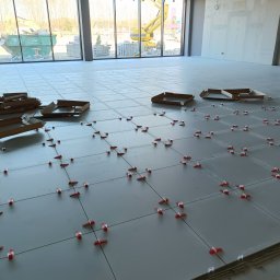 MIŁOSZ TOMKOWICZ - Układanie szarych płytek podłogowych z użyciem czerwonych klipsów poziomujących. W tle okno z widokiem na kontener i sprzęt budowlany. Kartony po płytkach na podłodze.