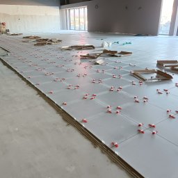 MIŁOSZ TOMKOWICZ - Układanie jasnych płytek podłogowych z użyciem czerwonych klipsów poziomujących w przestronnym, surowym wnętrzu. Widoczne narzędzia i kartony po płytkach.