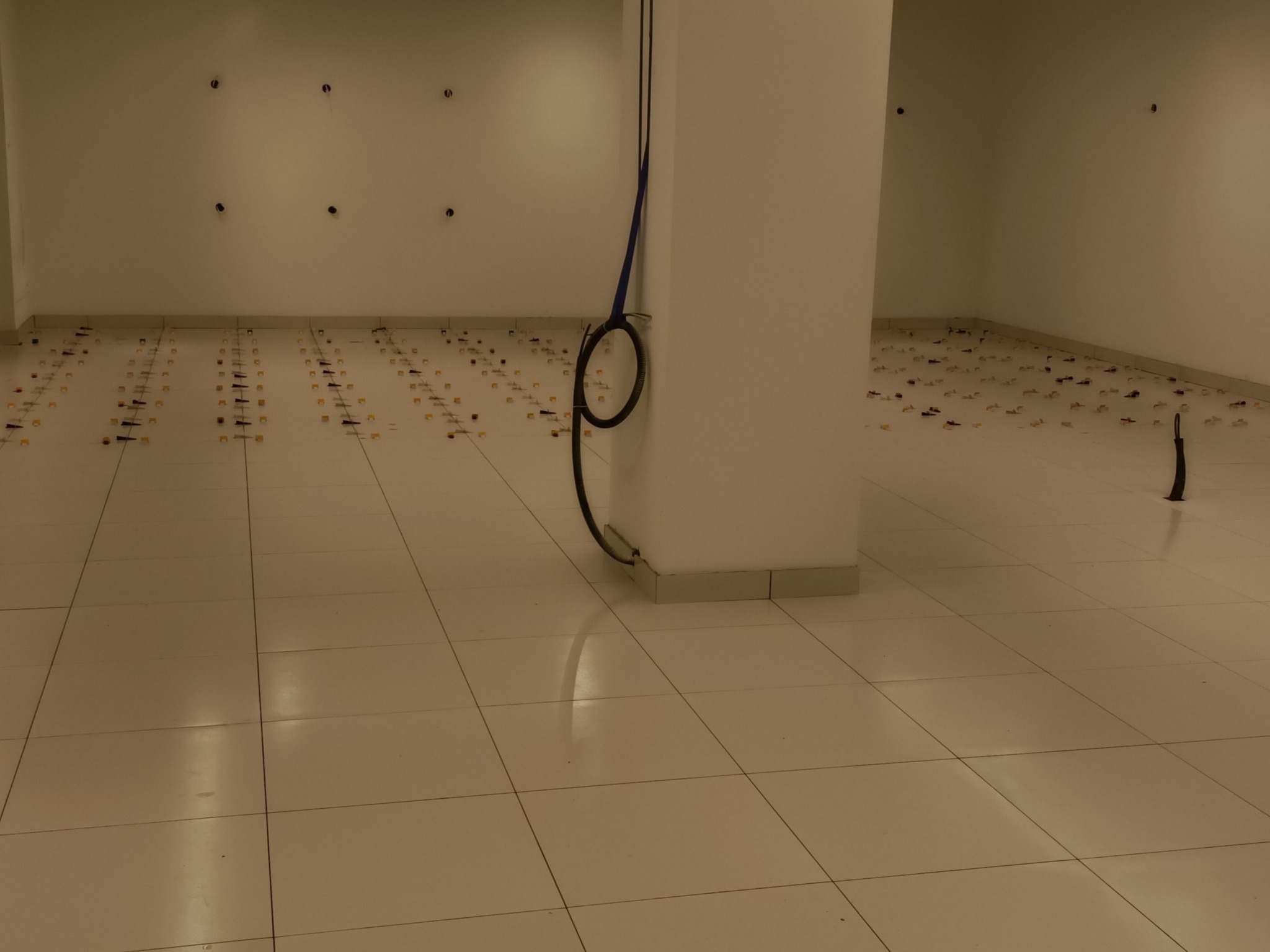 Przygotowanie do układania płytek podłogowych w dużym pomieszczeniu, widoczne krzyżyki dystansowe i poziomica oparta o filar, surowy stan, brak oświetlenia.