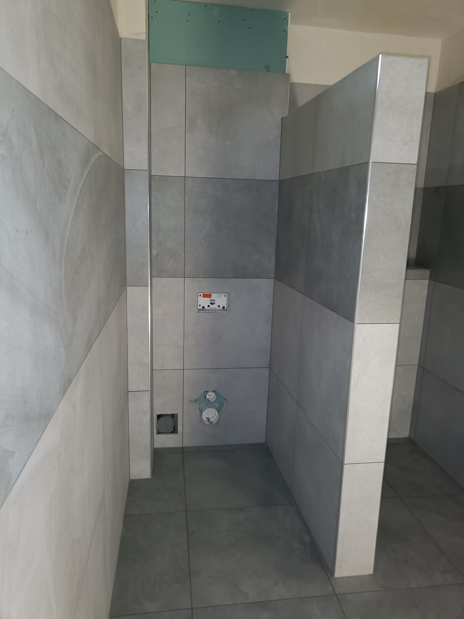 Wnętrze łazienki w trakcie układania szarych, prostokątnych płytek, widoczne podłączenia hydrauliczne i fragment płyty gipsowo-kartonowej w górnej części ściany.